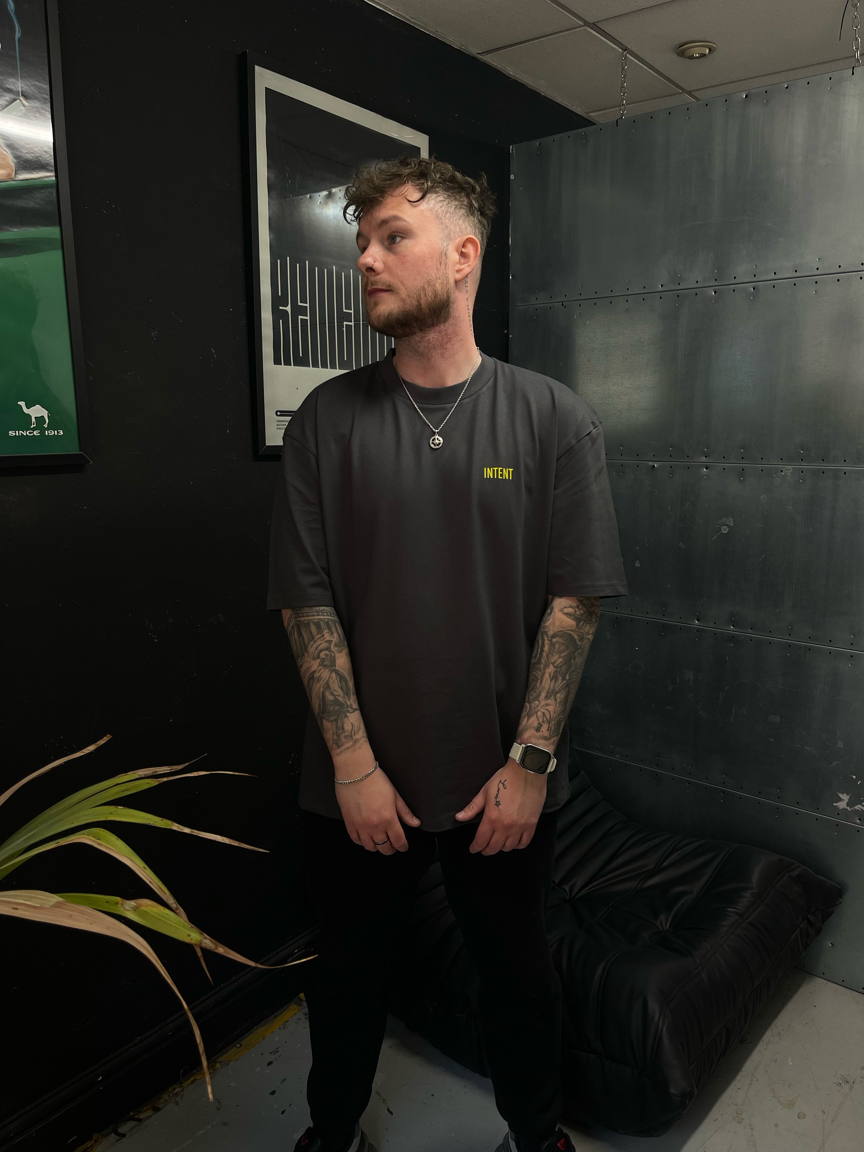 Man wearing a dark t-shirt with a logo in a room with a plant and framed pictures on the wall.