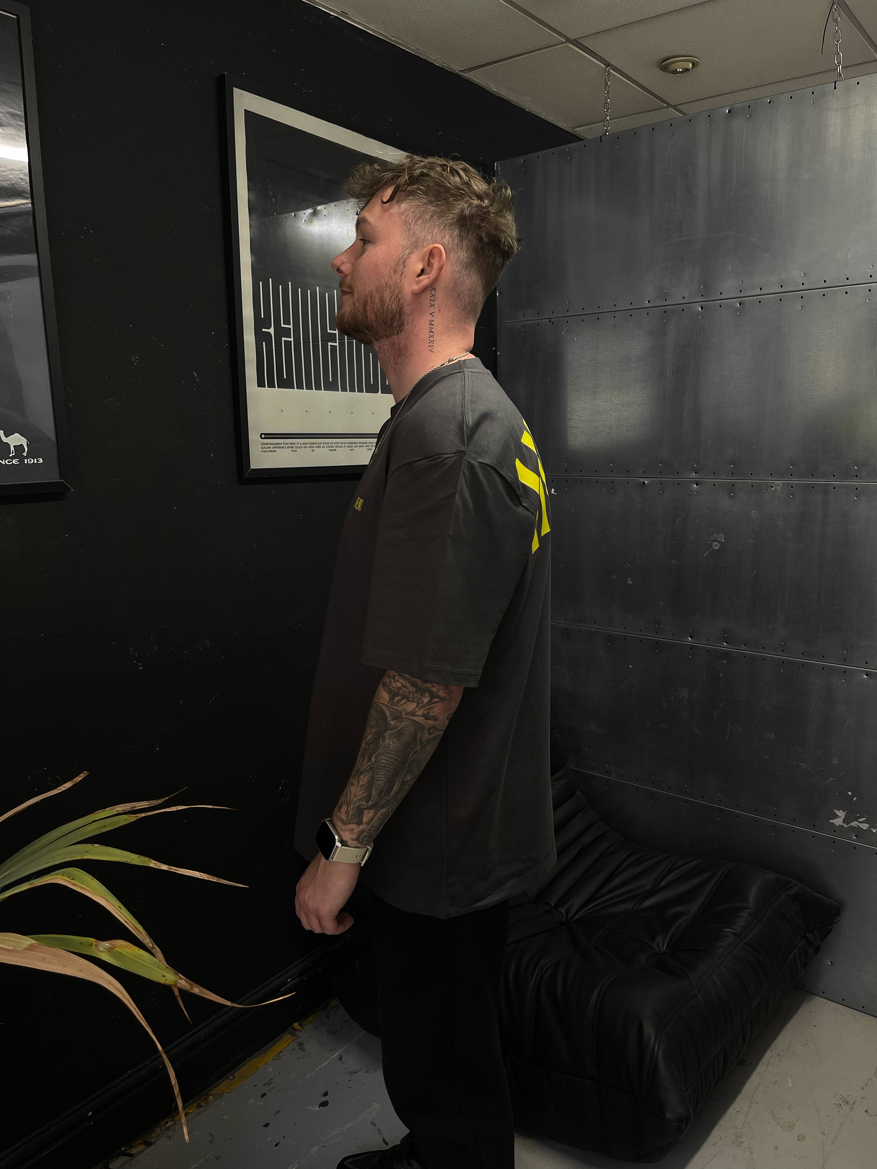 Man standing in a room with a black wall and framed picture, wearing a dark t-shirt with yellow accents.