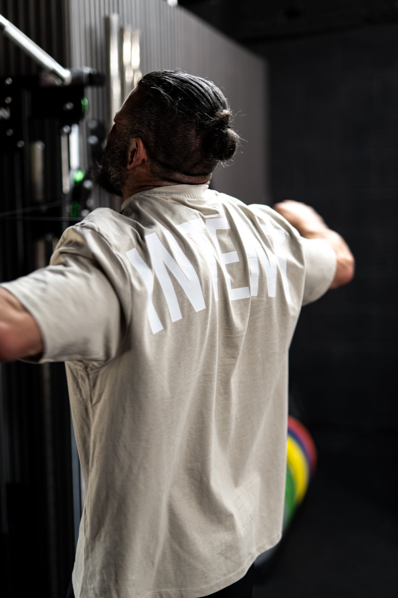 Person wearing a beige shirt with 'RIVER' printed on the back, standing in a room with dark walls and a colorful object in the background.