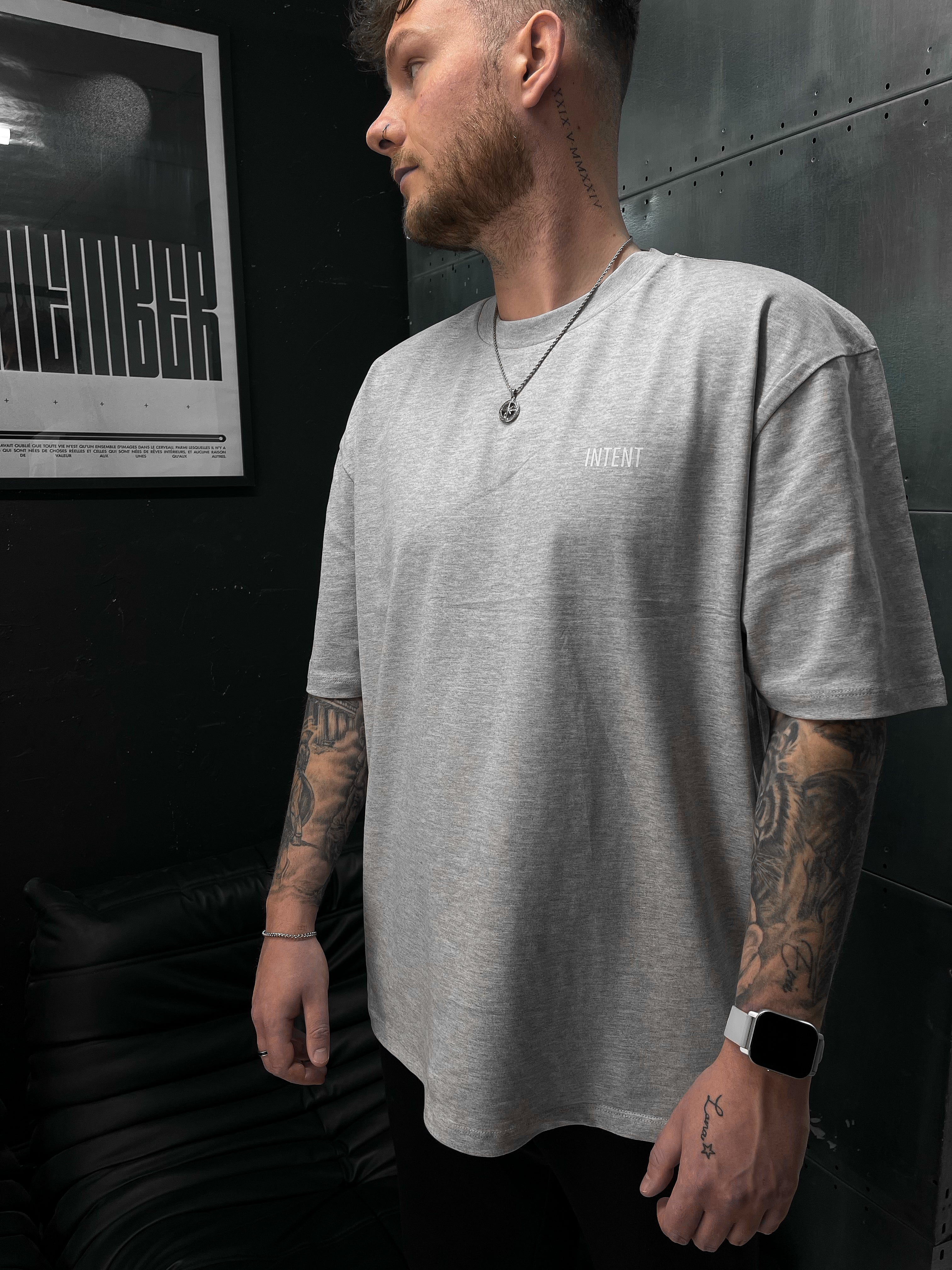 Man wearing a gray t-shirt with a logo, standing against a dark wall.