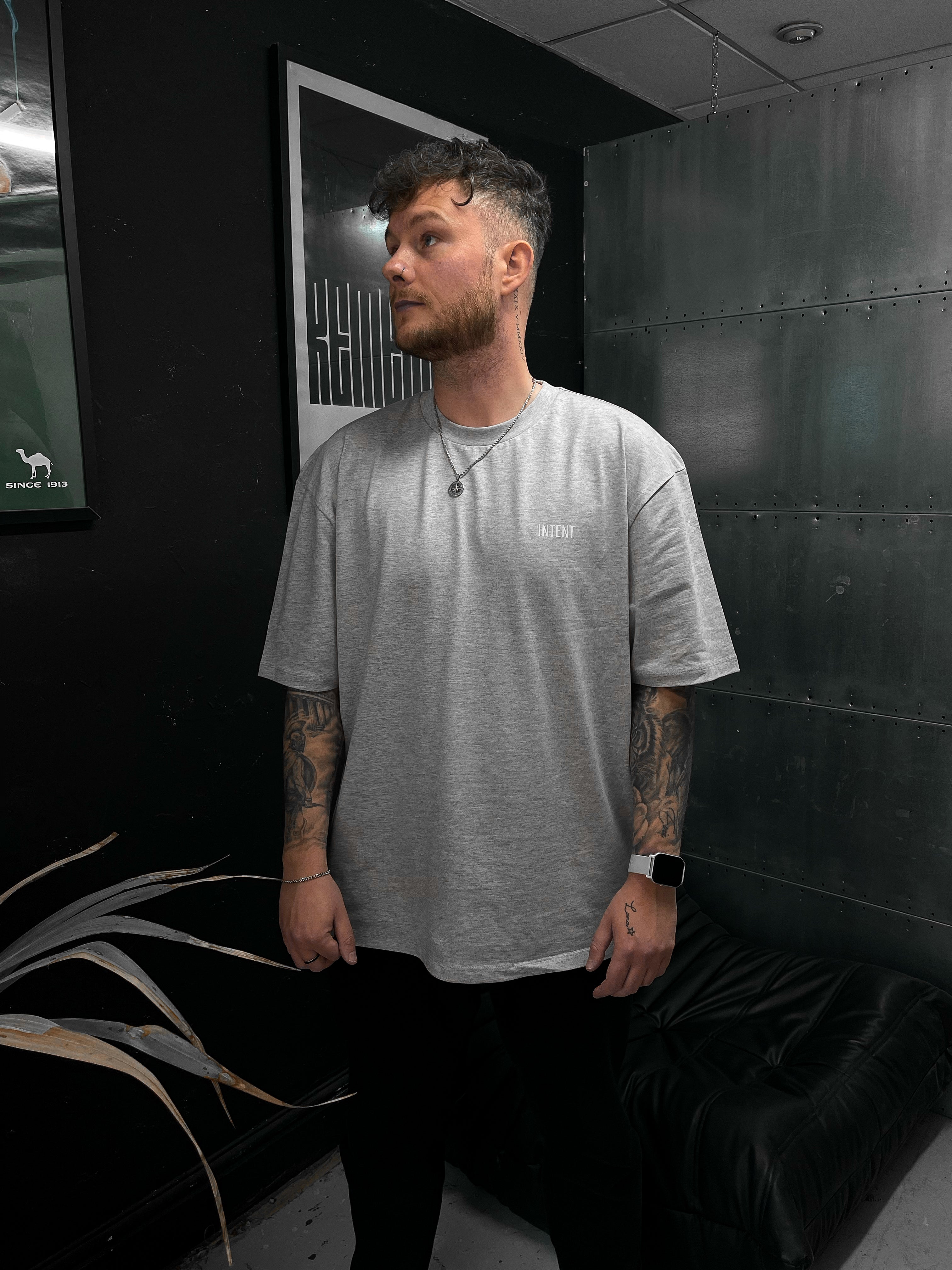 Man wearing a gray t-shirt in a room with dark walls and a plant.