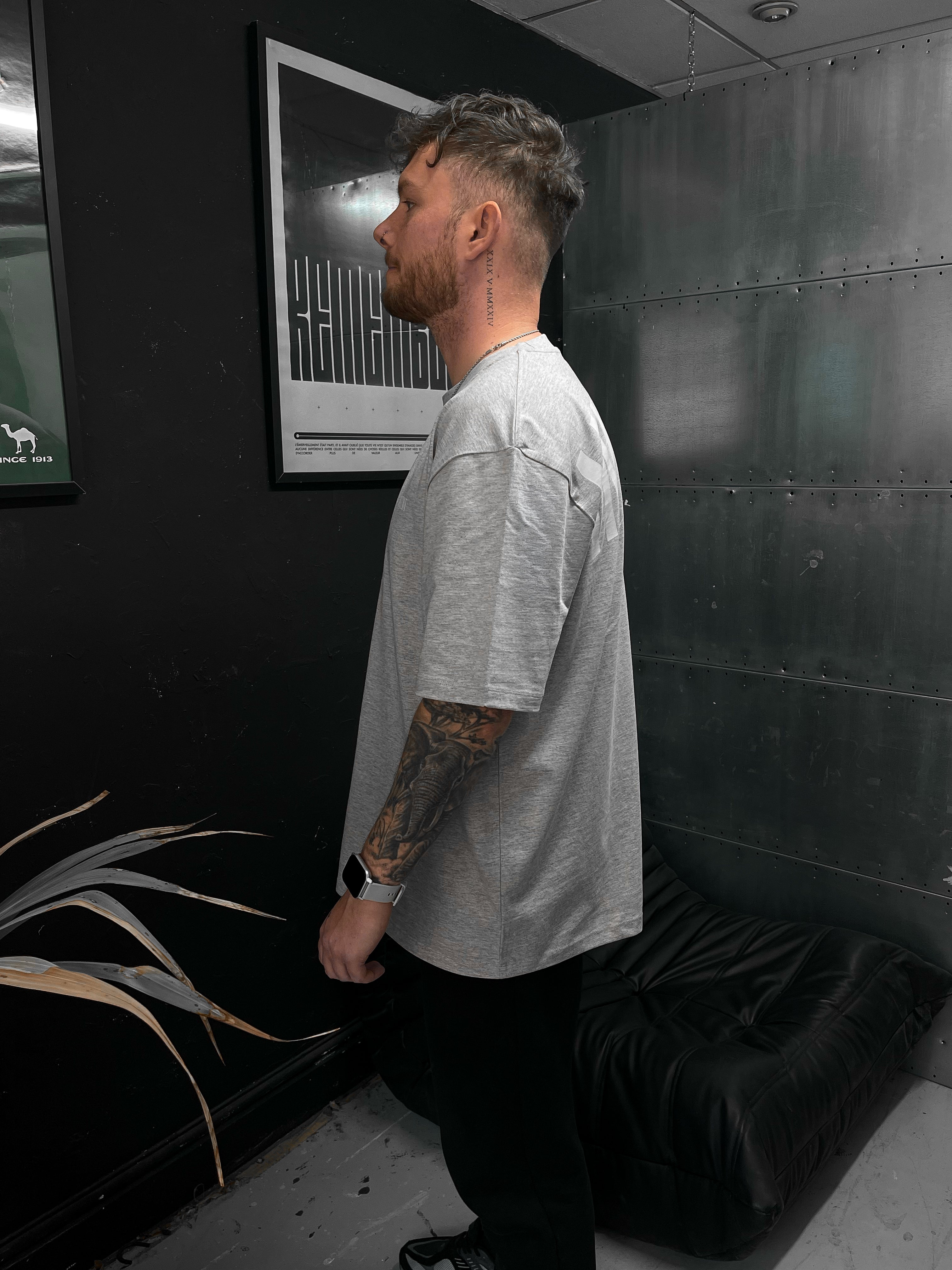 Man with tattoos standing in a room with dark walls and a plant.