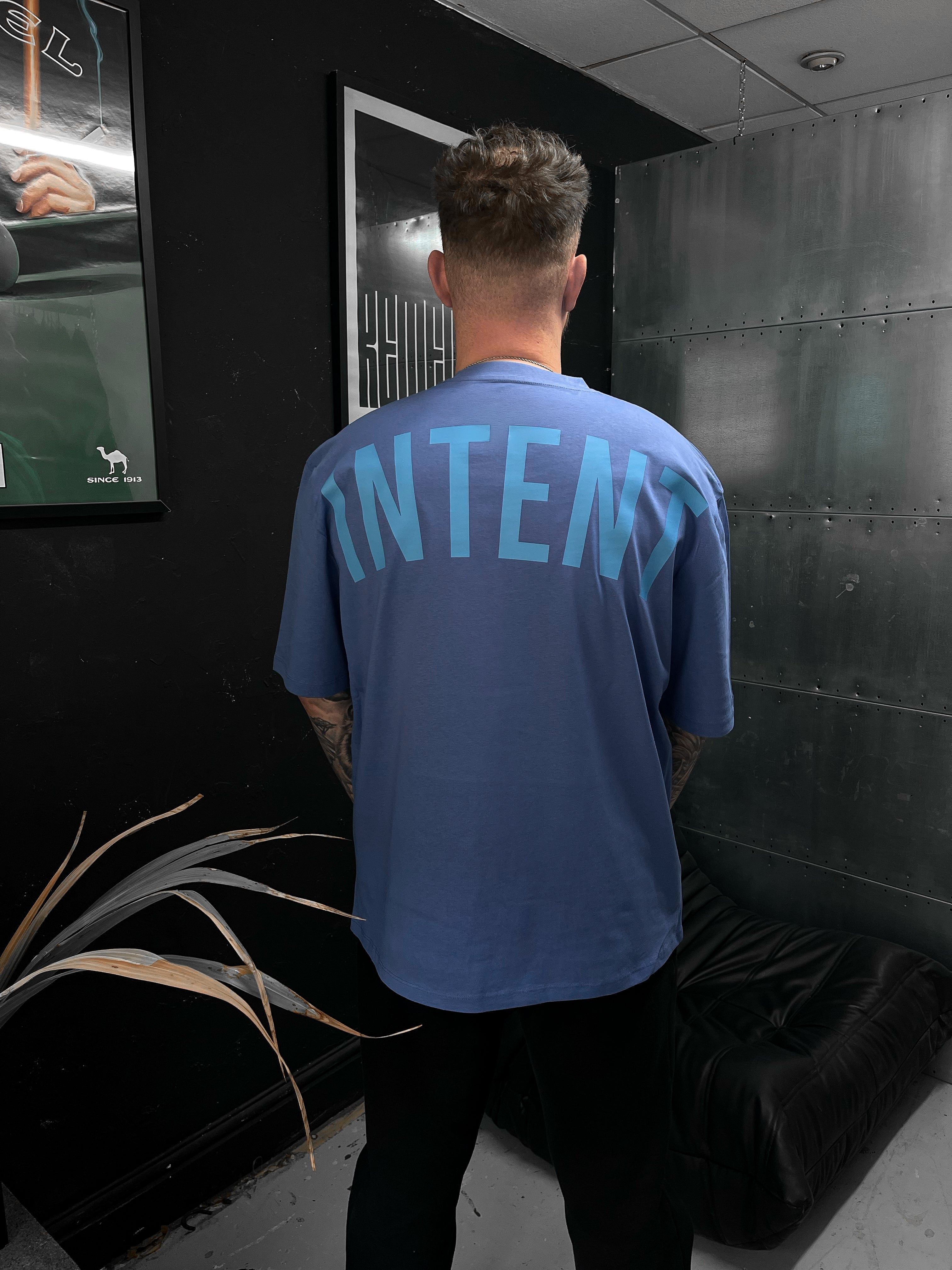 Person wearing a blue t-shirt with 'INTENT' printed on the back in a room with black walls and a plant.