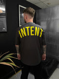 Person wearing a black t-shirt with 'INTENT' printed in yellow on the back, standing in a room with a plant and framed picture.