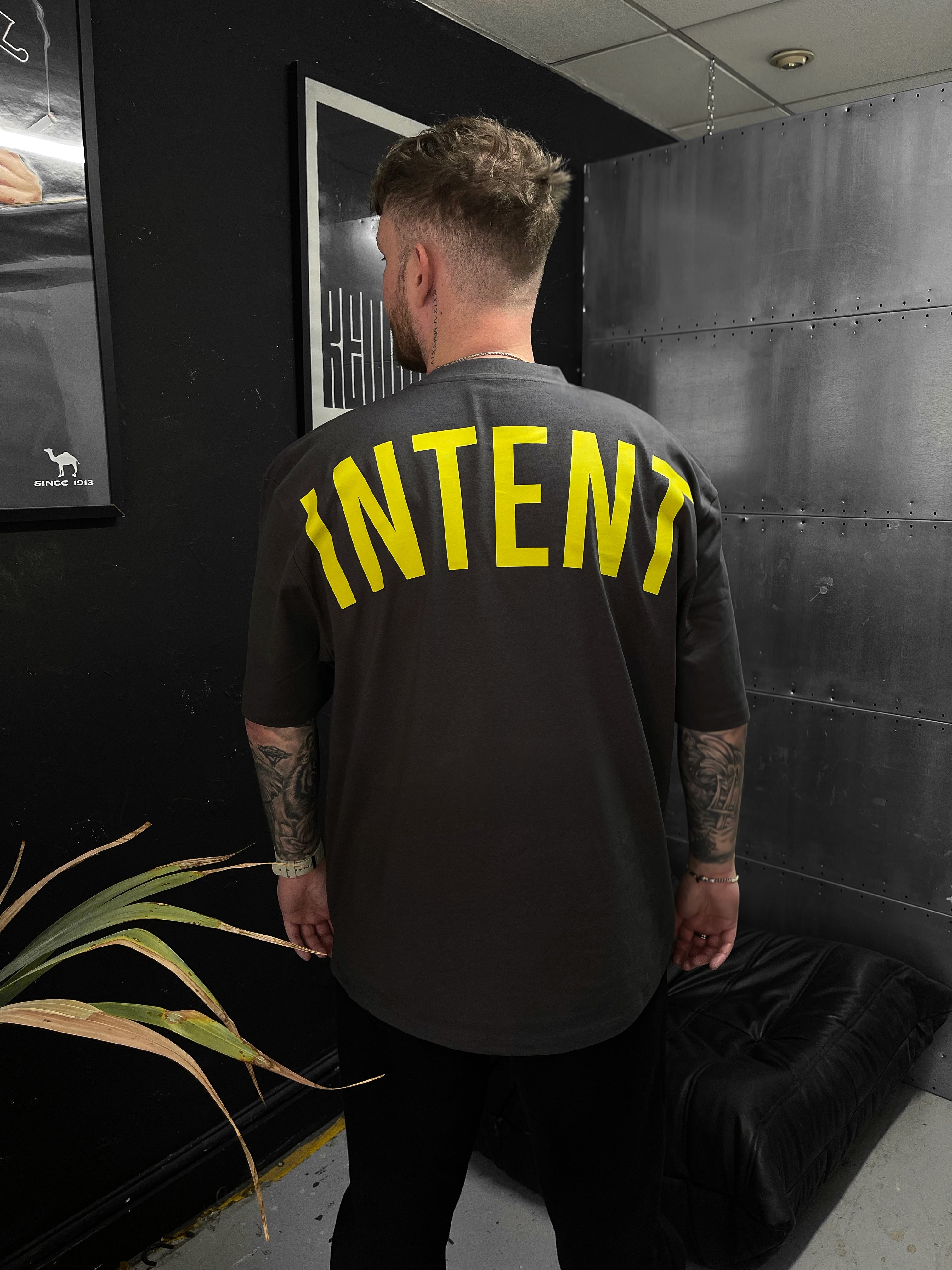 Person wearing a black t-shirt with 'INTENT' printed in yellow on the back, standing in a room with a plant and framed picture.