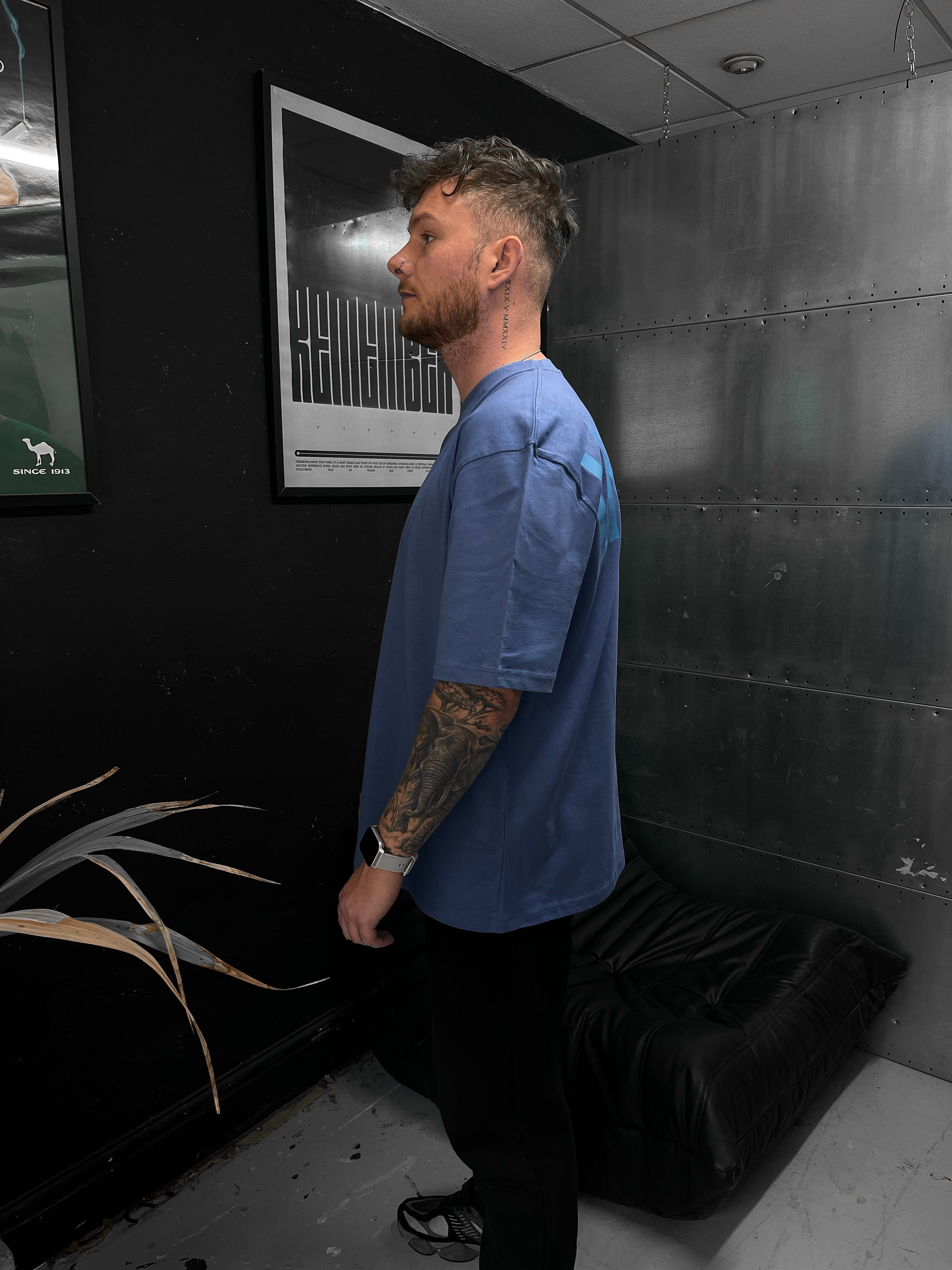 Man in a blue shirt and black pants standing in a room with dark walls and a plant.