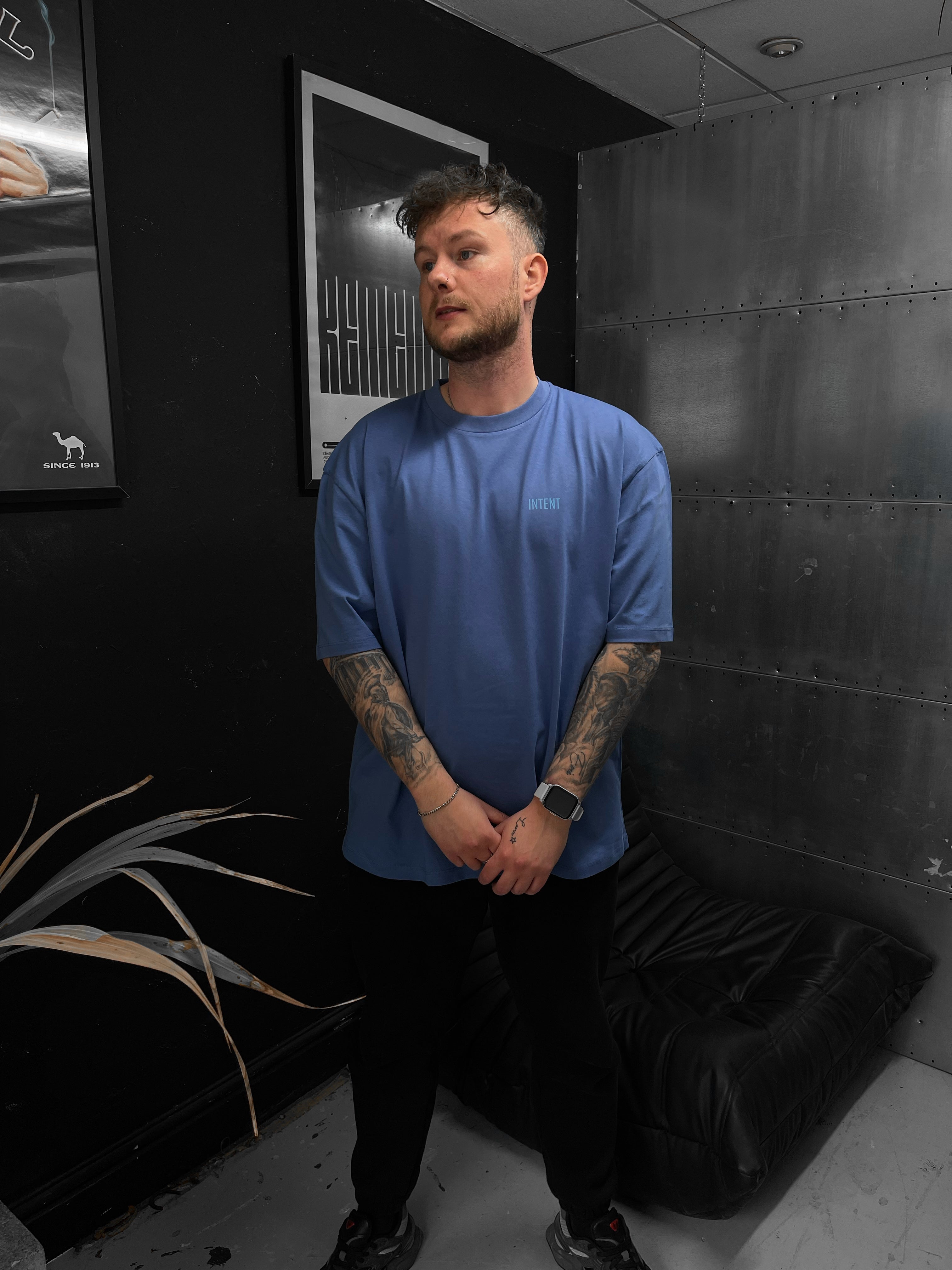 Man in a blue t-shirt standing in a room with dark walls and a black bag on the floor.
