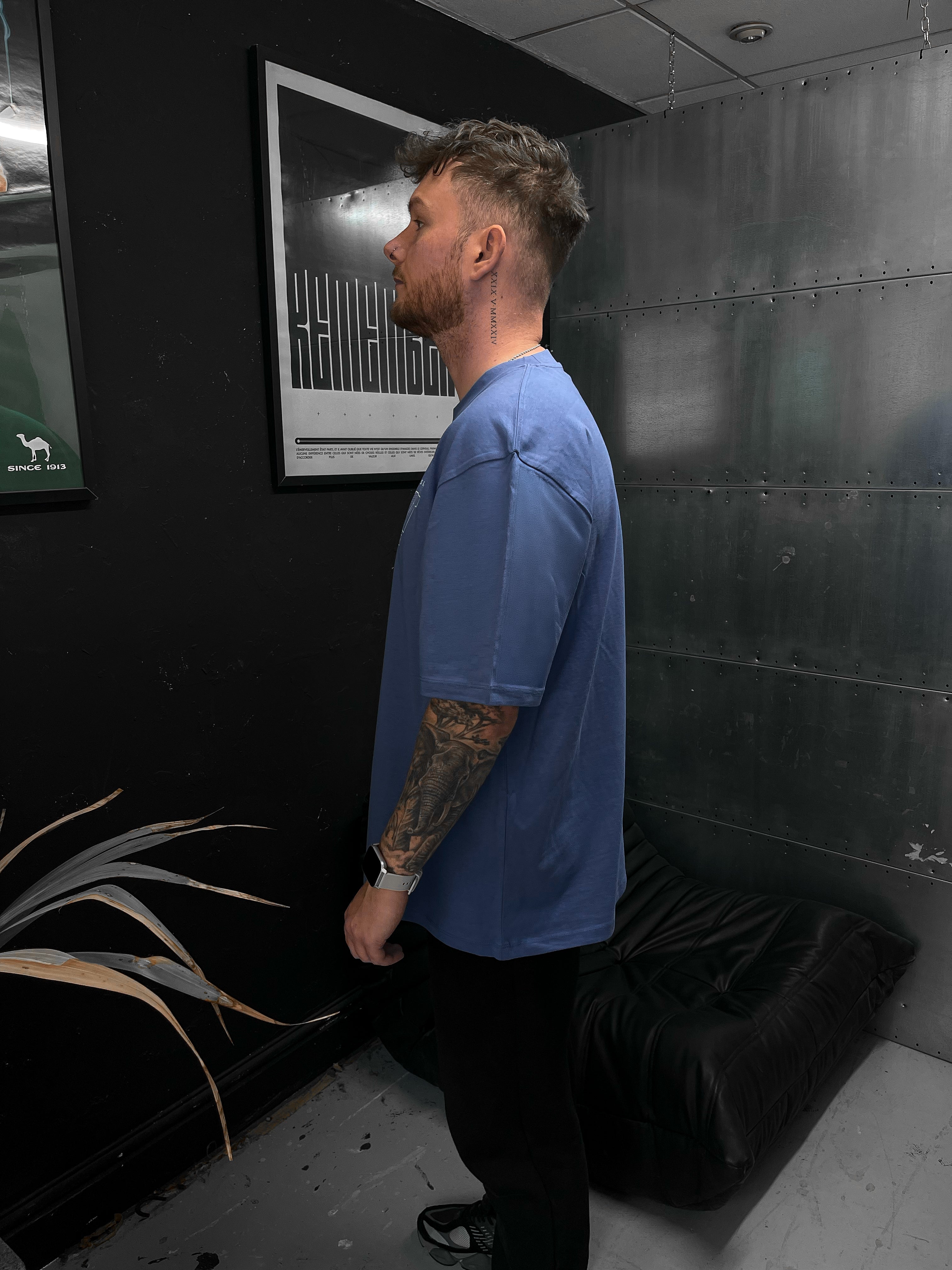 Man in a blue shirt standing in a room with a black wall and a plant.