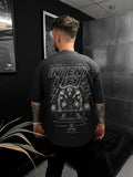 Person wearing a black t-shirt with graphic design and text, sitting in a room with dark walls and a plant.