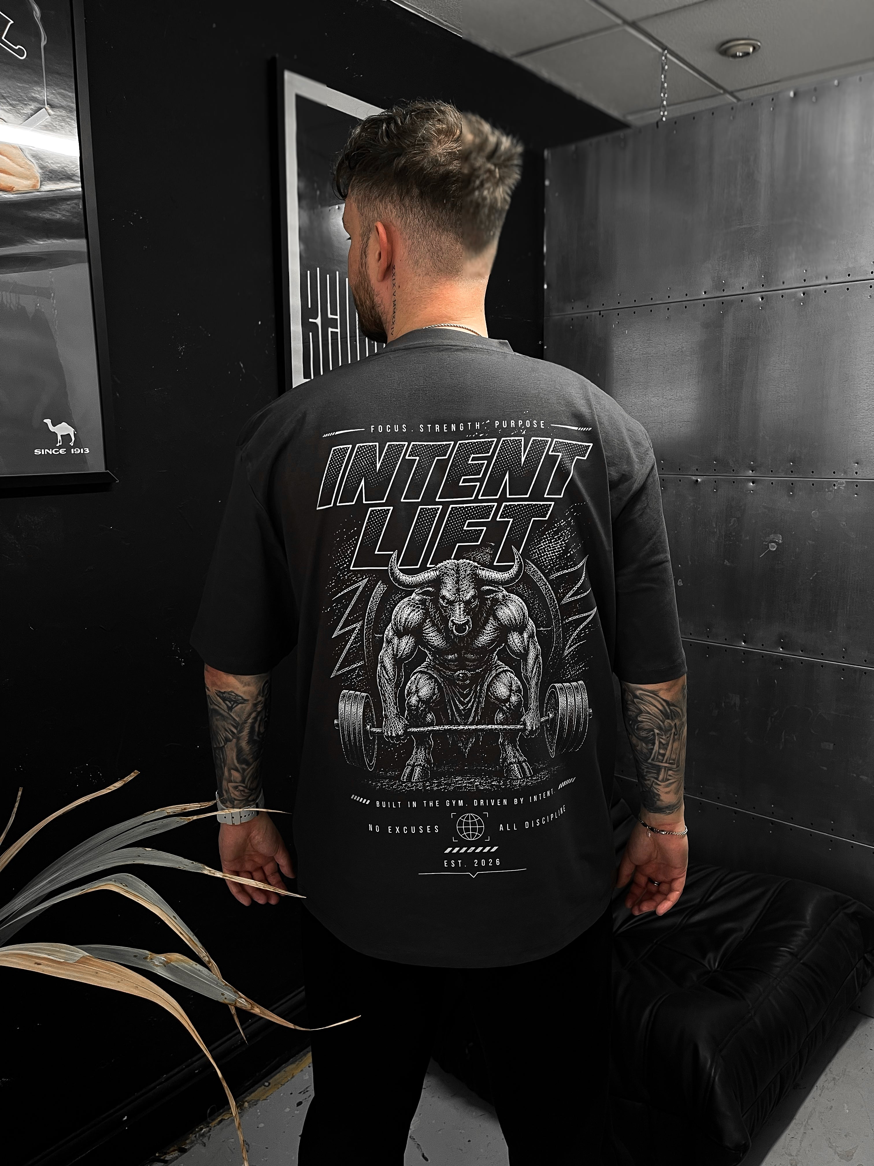 Person wearing a black t-shirt with graphic design and text, sitting in a room with dark walls and a plant.