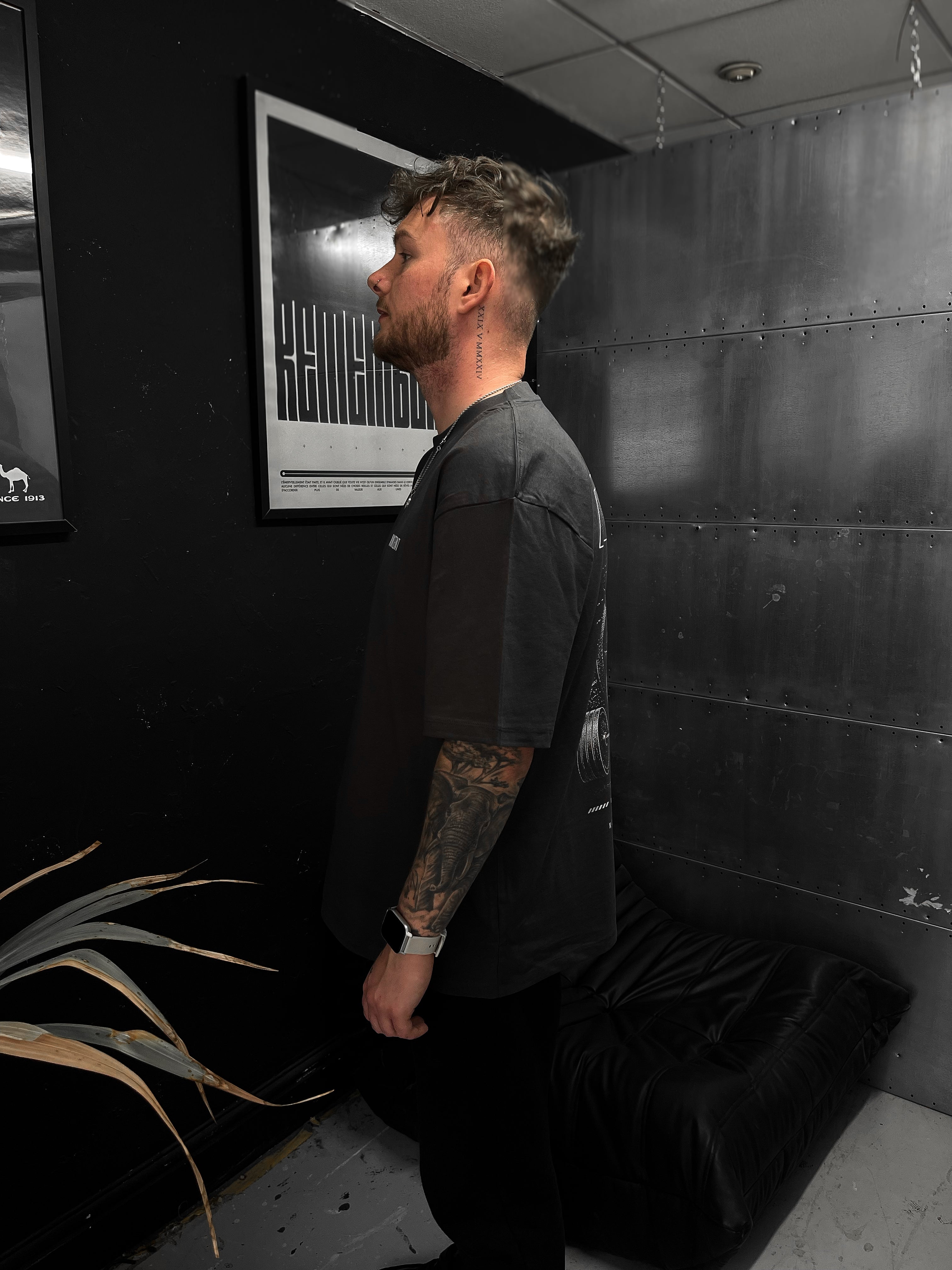 Man in a black t-shirt standing in a room with a plant and a wall with text.