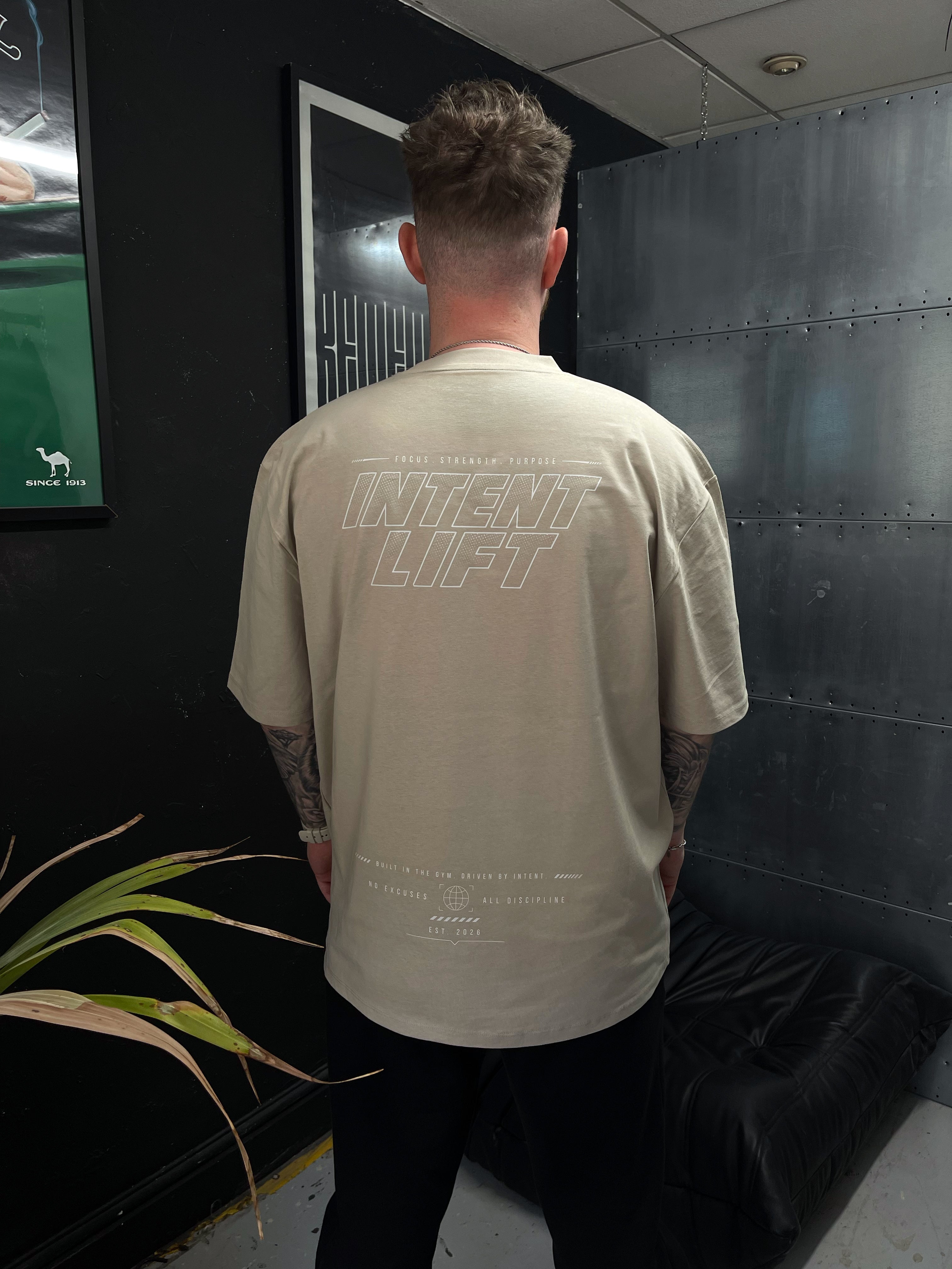 Person wearing a beige t-shirt with 'Intent Lift' text on the back, standing in a room with dark walls and a plant.