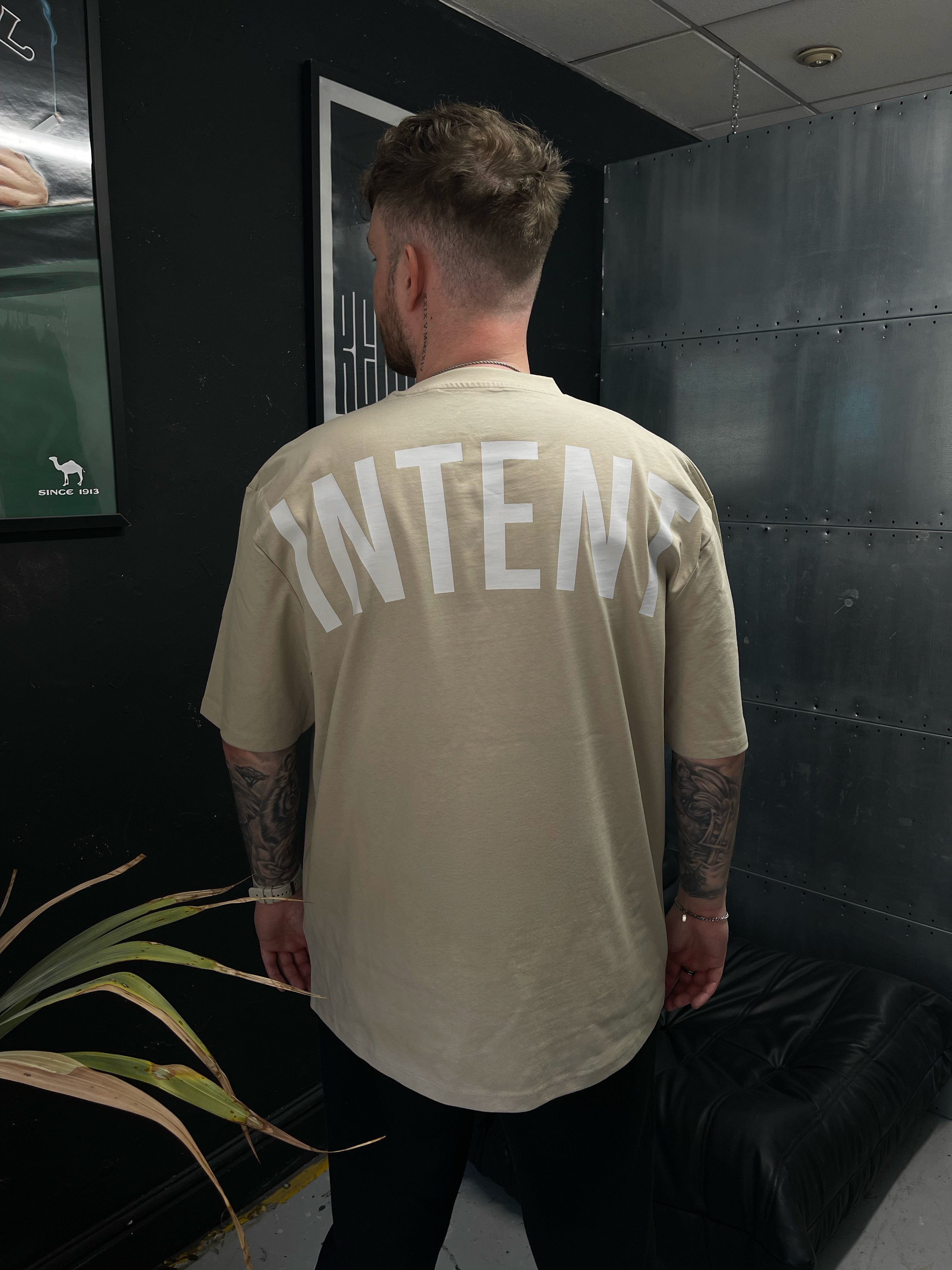 Person wearing a beige t-shirt with 'INTENT' printed on the back in a dark indoor setting.