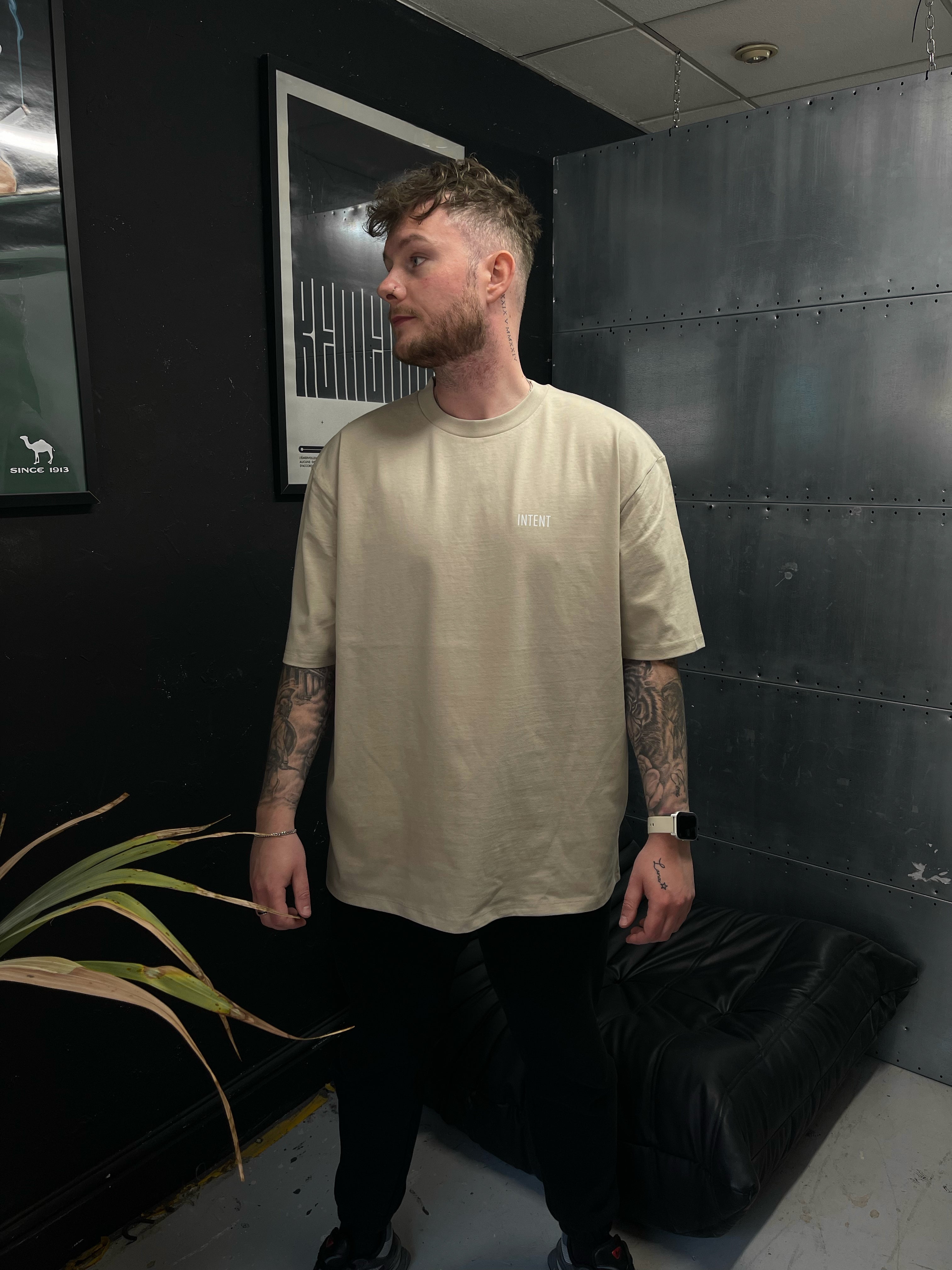 Man wearing a beige t-shirt standing in a room with dark walls and a plant.